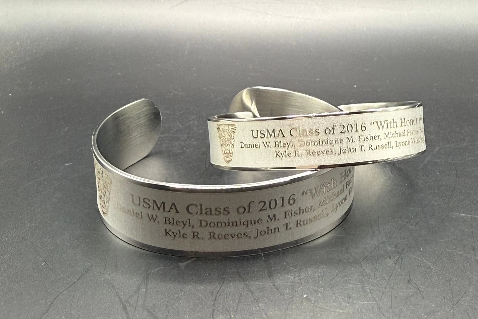 Two stainless-steel USMA Class of 2016 memorial bracelets showing the class title and engraved seven-name roster.